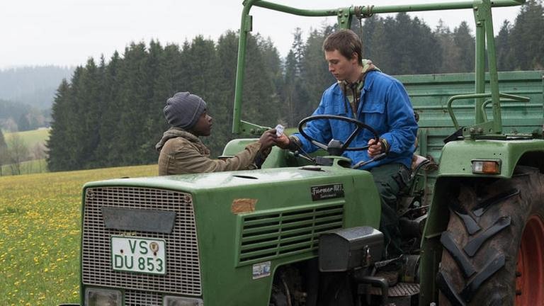 Tayo zeigt einem jungen Mann auf einem Traktor einen Zettel
