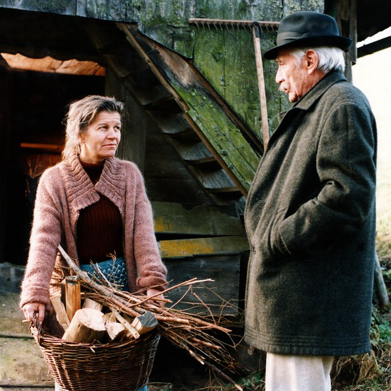 Lioba trägt einen Korb mit Holz und spricht mit Wilhelm
