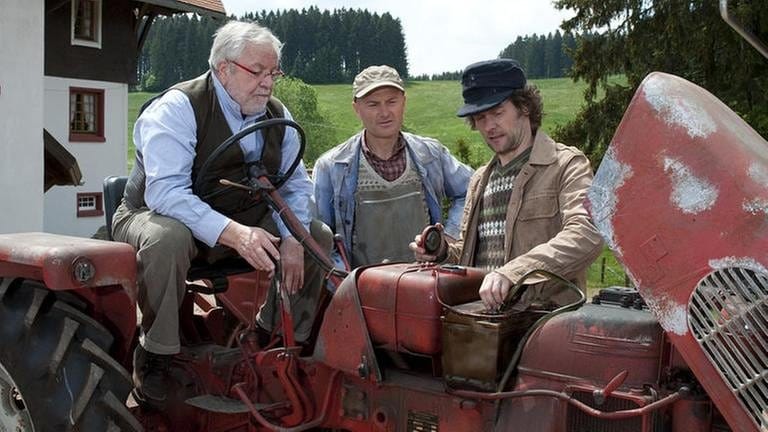 Hermann sitzt auf einem alten roten Traktor, Karl und Riedle stehen bei daneben