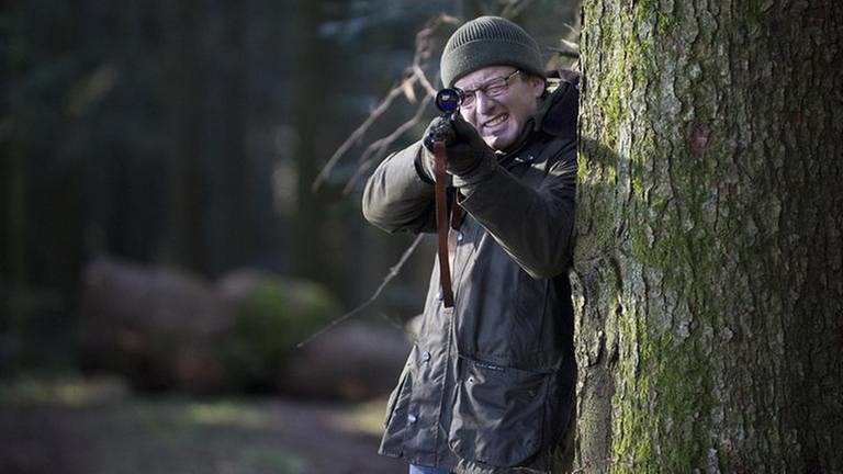 Bernhard steht in seinem Wald an einen Baum gelehnt und legt die Flinte an