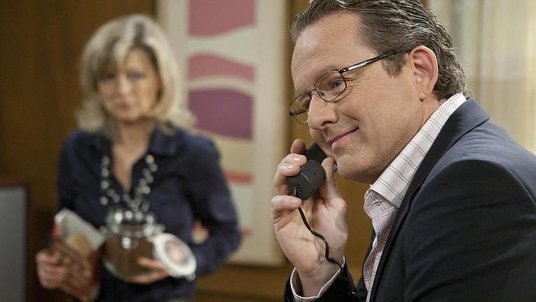 Bürgermeister Faller telefoniert in seinem Büro, im Hintergrund sieht man schemenhaft seine Sekretärin, Frau Heilert