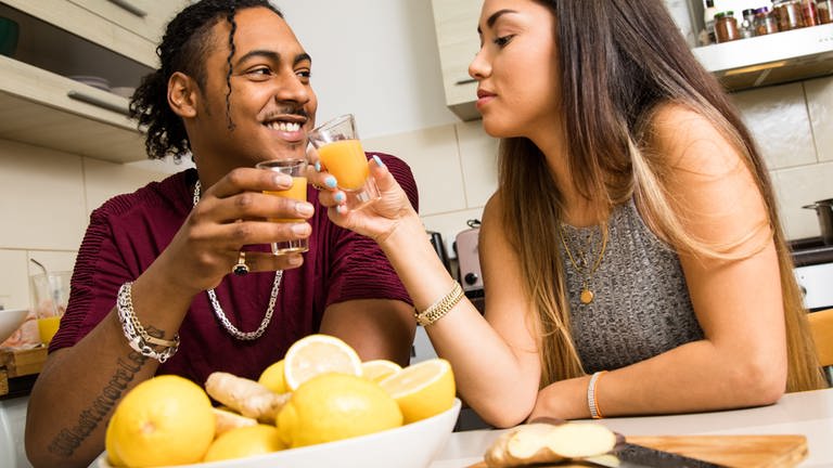 Ein Paar hält am Küchentisch Ingwer-Shots mit Zitrone und Apfel in den Haenden und prostet sich zu.