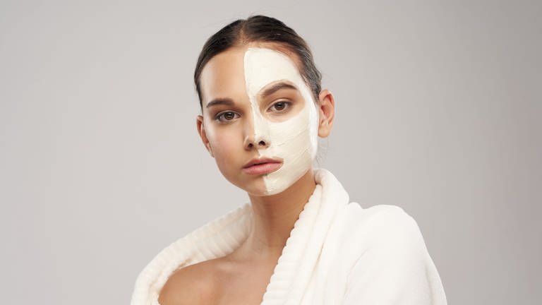 Junge Frau mit neutralem Blick und kosmetischer Creme-Gesichtsmaske, die nur auf einer Gesichtshälfte aufgetragen ist. 