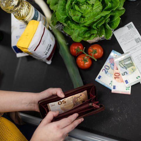 Person öffnet an einer Kasse ihren Geldbeutel. Auf dem Kassenband liegen verschiedene Lebensmittel, drei Euro-Scheine und der Kassenzettel.