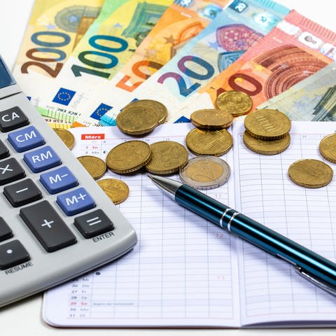 In der linken Bildhälfte liegt ein Taschenrechner. In der Mitte diverse Euro-Scheine und -münzen, sowie ein Kalendarplaner und ein schwarzer Kugelschreiber.