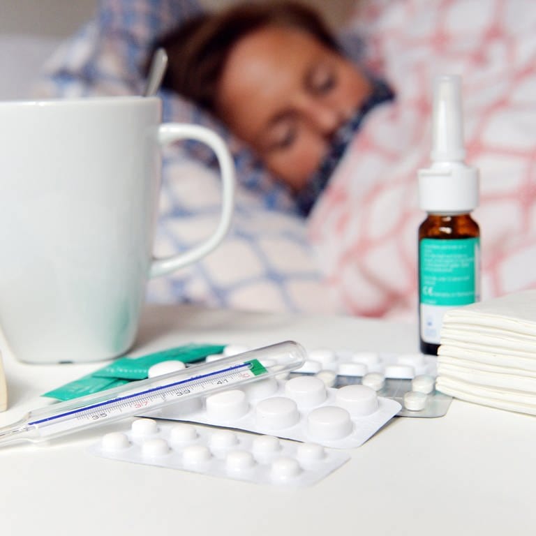 Frau liegt krank im Bett, davor Medikamente, Tee, Nasenspray und Fieberthermometer auf dem Nachttisch. Was hilft bei akuter und chronischer Rhinosinusitis? 