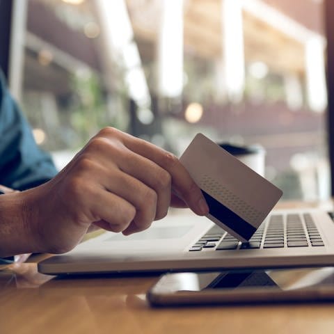 Ein Mann hält in seiner rechten Hand eine Kreditkarte. Diese ist auf die Tastatur eines Laptops angelehnt, welcher vor ihm steht. Auf der rechten Seite des Laptops liegt ein Smartphone.