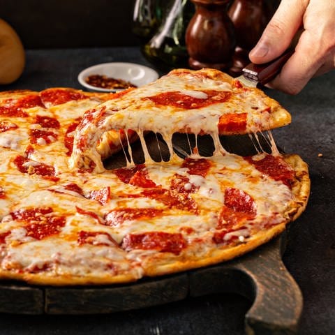 Eine Hand hebt ein Stück einer vorgeschnittenen Salami Fertigpizza von einem Holzbrett. Der Käse zieht Fäden.