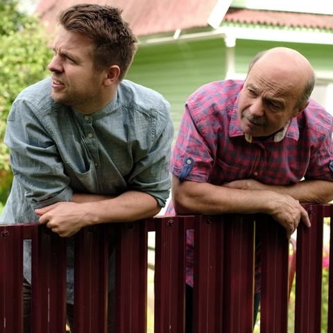 Zwei Männer mittleren Alters lehnen sich stirnrunzelnd über einen Gartenzaun und schauen auf das Nachbargrundstück.