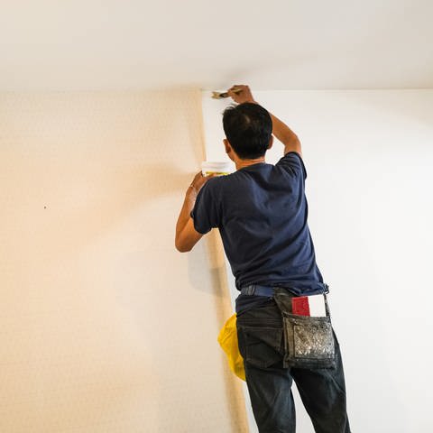 Ein Handwerker klebt mit einem Pinsel Tapete auf eine Wand.