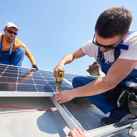 Zwei Handwerker bringen auf einem Dach Solarmodule einer Photovoltaikanlage an. Mit einem Akkuschrauber wird das Solarpanel an das Dach befestigt.
