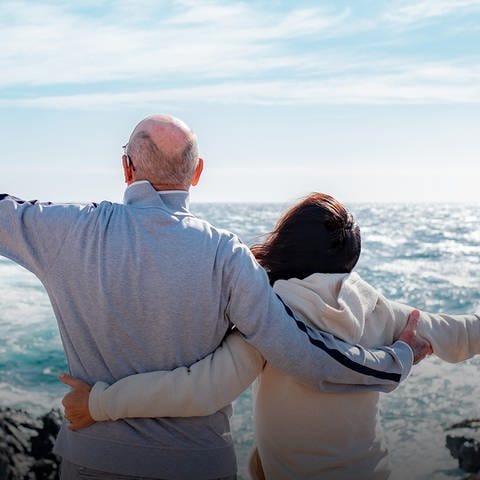 Im Hintergrund ist ein Meer zu sehen, im Vordergrund ist ein Rentner-Paar von hinten zu sehen, das sich im Arm hält und jeweils einen Arm in Richtung Horizont streckt.