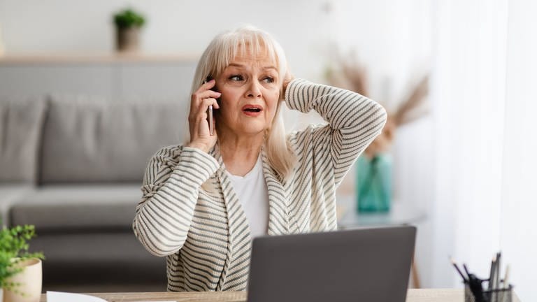 Geschockte ältere Dame telefoniert vor ihrem Laptop im Wohnzimmer. (Foto: Adobe Stock, Adobe Stock/Prostock-studio)