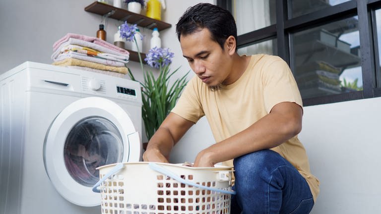 Junger Mann befüllt eine Waschmaschine mit Wäsche. Was sagt die Füllmenge bei der Waschmaschine aus? Welche ist wirklich effizient?
