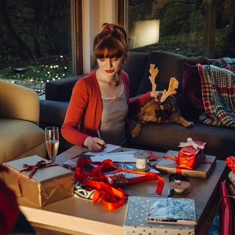 Junge Frau schreibt an Couchtisch Weihnachtspost, daneben ein Glas Sekt und eingepackte Geschenke. Wie kann KI dabei helfen, Geschenke zu finden?