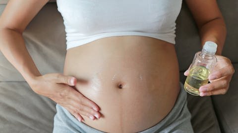 Schwangere Frau cremt sich mit Öl ihren Schwangerschaftsbauch ein. Sie liegt auf einem Bett. Die Frau trägt ein weißes bauchfreies Top und eine graue Hose.