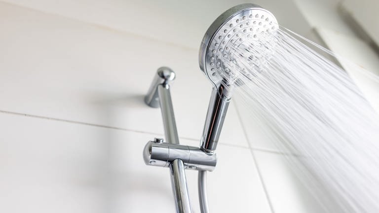 In einer Dusche hängt ein silbener Sparduschkopf. Die Dusche ist an und das Wasser schießt aus den Drüsen, obwohl ein wassersparender Duschkopf weniger Wasser pro Minute durchlässt.