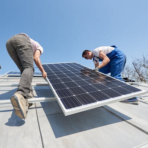 Zwei Männer installieren ein Panel einer Solaranlage auf einem Hausdach. 